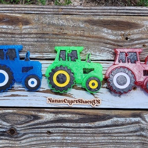 May include: Three toy tractors, one blue, one green, and one red, are arranged in a row on a wooden surface. Each tractor has black wheels with white rims and a black cab.
