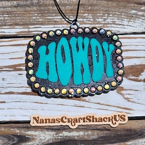 May include: A rectangular black pendant with a textured surface featuring the word HOWDY in bright teal letters. The border is lined with iridescent rhinestones. A black cord is attached to the top. A wooden sign below reads NanasCraftShackUS.