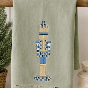 May include: A sage green hand towel featuring an embroidered nutcracker design in shades of blue, gold, and white. The nutcracker figure is centered on the fabric, which has a hemstitched border detail along the bottom edge of the towel.