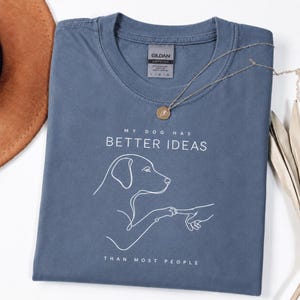 以下が含まれることがあります： スレートブルーのTシャツで、犬が人の指に触れている白い線画が描かれています。テキストは「MY DOG HAS BETTER IDEAS THAN MOST PEOPLE」と書かれています。Tシャツの首元にはGildan Softstyleのラベルが付いています。