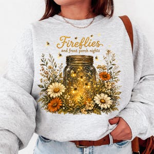May include: A light grey sweatshirt with a mason jar design filled with glowing lights and flowers. The text "Fireflies and front porch nights" is printed above the jar in gold lettering.