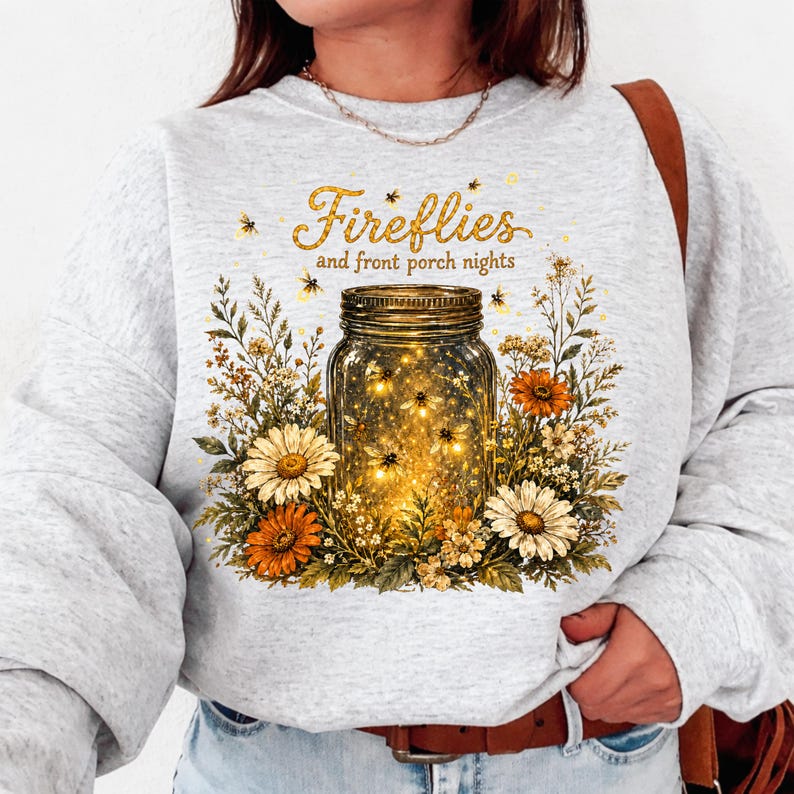 May include: A light grey sweatshirt with a mason jar design filled with glowing lights and flowers. The text "Fireflies and front porch nights" is printed above the jar in gold lettering.