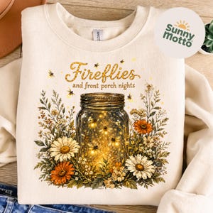 May include: A cream-coloured sweatshirt with a mason jar design filled with glowing fireflies, surrounded by orange, white, and green floral elements. The text "Fireflies and front porch nights" is arched above the jar.