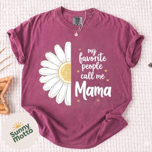 Peut inclure: T-shirt de couleur bordeaux avec une marguerite blanche et jaune. Le texte "my favorite people call me Mama" est imprimé en blanc, avec de petits cœurs dorés. Un vêtement décontracté.