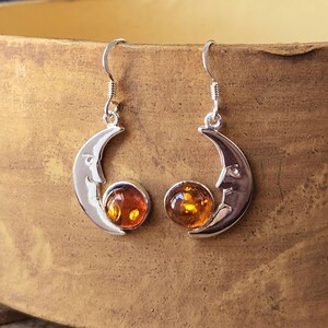 May include: A pair of silver crescent moon earrings with amber gemstones. The earrings are hanging from a wooden surface.