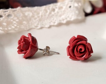 May include: A pair of red rose stud earrings. The earrings are made of a red material that looks like coral. The earrings are on a white surface.