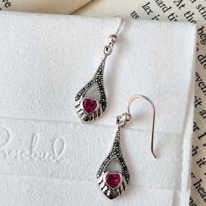May include: A pair of silver dangle earrings with a teardrop design. Each earring features a small red heart-shaped gemstone in the center.