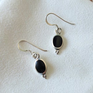 May include: A pair of silver earrings with a black oval stone. The earrings have a hook design and a small decorative element at the bottom. The earrings are displayed on a white cloth.