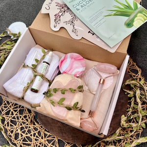May include: A white gift box filled with a variety of skincare products, including a face mask, a jade roller, a gua sha tool, and a bottle of serum. The box is decorated with green leaves and twine.