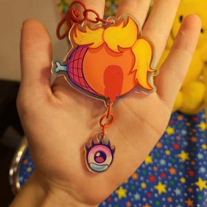 May include: A clear acrylic keychain featuring a large orange ham-shaped graphic with yellow hair and a red drip, attached to a smaller dangling charm of a stylized purple and pink eye with a flame-like border, connected by a red metal clasp.