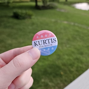 May include: A round campaign button with a red, white, and blue color scheme. The button features the name "KURTIS" in bold white letters, with "FOR MAYOR" below. The top half has red stripes and white stars, and the bottom half has blue stripes and white stars.
