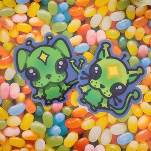 May include: Two green alien creature stickers with cartoon eyes and a blue background. The stickers are on a background of colourful jelly beans.