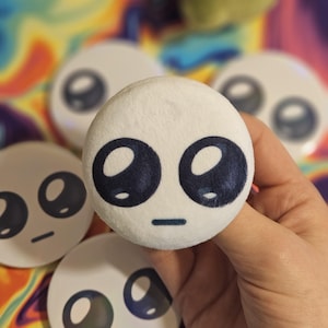 Puede incluir: Un objeto redondo y blanco de felpa con una cara de dibujos animados. Presenta ojos grandes de color azul oscuro y una pequeña línea horizontal para la boca. Varios objetos similares son visibles en el fondo.