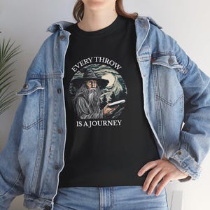 Puede incluir: Camiseta negra con un gráfico de mago y el texto "EVERY THROW IS A JOURNEY". El mago sostiene un disco, con un fondo de montaña y luna. La camiseta está hecha de un material suave.