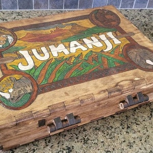 May include: A wooden game box with a painted design featuring the word "JUMANJI" and jungle animals. The box has a hinged lid and metal clasps.