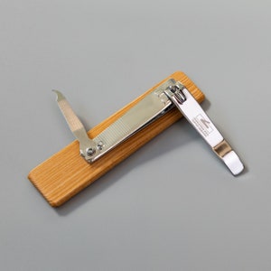 May include: A silver nail clipper with a wooden handle. The clipper has a file and a nail cleaner attached to it. The handle is made of light brown wood. The clipper has the text "CHARACTERISTIC ART CLIPPER WORKS" engraved on the side.