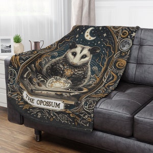 May include: A black and gold throw blanket featuring a whimsical illustration of an opossum holding a glowing orb. The text "The Opossum" is written in a decorative font on the blanket.