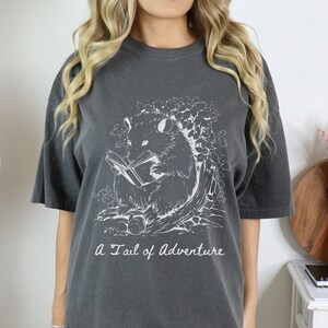 Puede incluir: Una camiseta gris oscuro con un gr&aacute;fico blanco de un opossum leyendo un libro. El texto "A Tail of Adventure" est&aacute; impreso debajo del gr&aacute;fico.