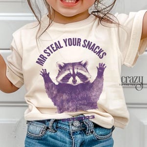 Pu&ograve; includere: T-shirt color crema con un'illustrazione di procione viola e la scritta "MR STEAL YOUR SNACKS". Il procione ha le braccia alzate. La maglietta ha anche la scritta "crazy to the tee" e il numero "123".