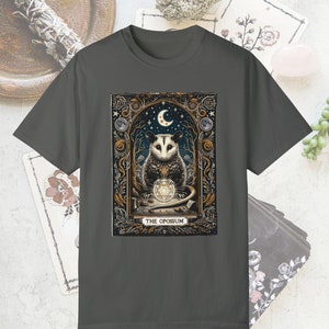 May include: A dark gray t-shirt with a graphic print of a possum holding a crystal ball. The graphic is surrounded by a gold frame and features a crescent moon and stars. The text "THE OPOSSUM" is printed below the graphic.