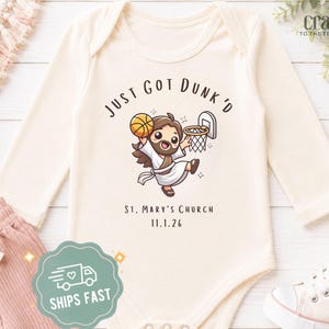 May include: A cream-colored long-sleeve baby bodysuit featuring a cartoon illustration of a figure playing basketball with the text "JUST GOT DUNK'D" above and "ST. MARY'S CHURCH 11.1.26" below. The garment has a round neckline and snap closures.