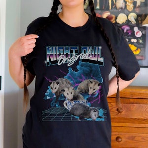 May include: Black t-shirt with a graphic of four opossums in a teacup. The graphic is in a retro style with neon colors and lightning bolts. The text "NIGHT OWL Original" is above the graphic.