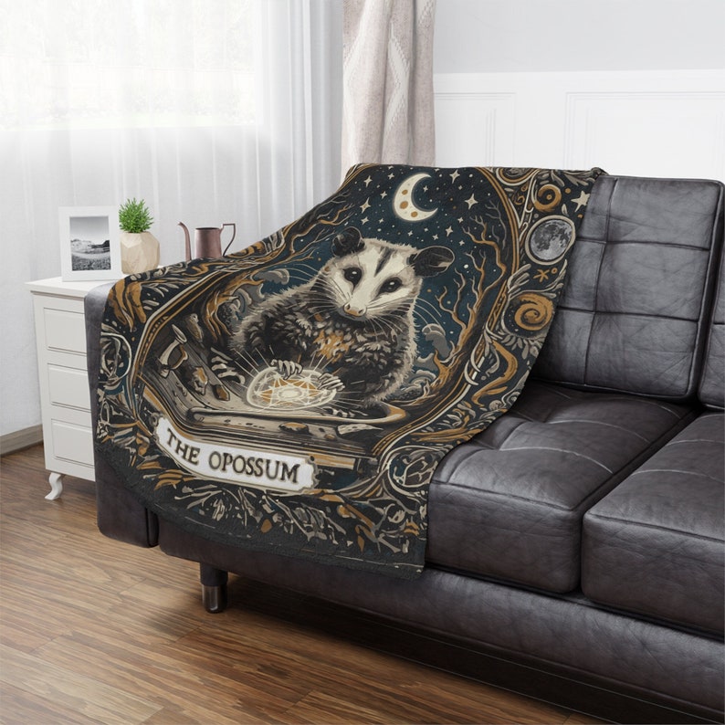 May include: A black and gold throw blanket featuring a whimsical illustration of an opossum holding a glowing orb. The text "The Opossum" is written in a decorative font on the blanket.