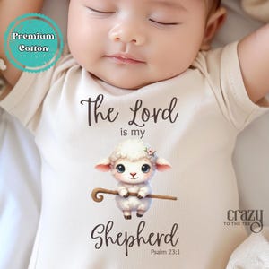 May include: A white baby onesie with a brown graphic of a lamb holding a shepherd's staff. The text "The Lord is my Shepherd" and "Psalm 23:1" is printed on the onesie.