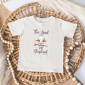 May include: A white baby onesie with a cute cartoon lamb holding a shepherd's staff. The text on the onesie reads "The Lord is my Shepherd, Psalm 23:1".