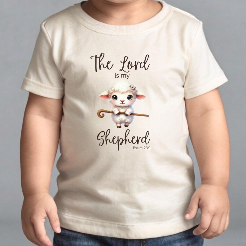 May include: A white toddler t-shirt with a cute cartoon lamb holding a shepherd's staff. The text "The Lord is my Shepherd" is printed on the shirt, along with the Bible verse "Psalm 23:1".