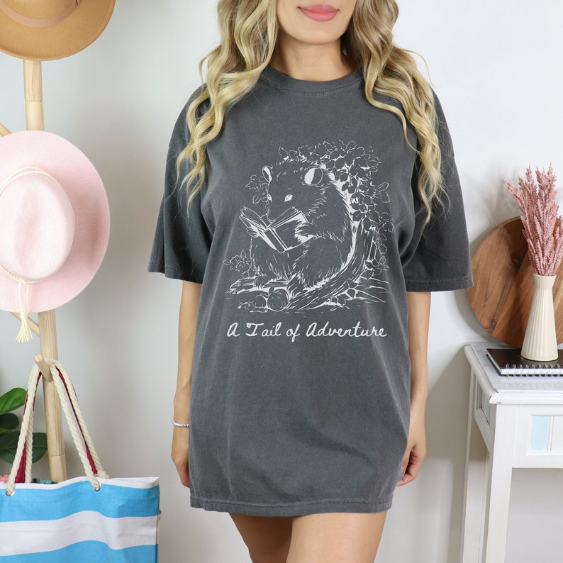 Puede incluir: Una camiseta gris oscuro con un gr&aacute;fico blanco de un opossum leyendo un libro. El texto "A Tail of Adventure" est&aacute; impreso debajo del gr&aacute;fico.