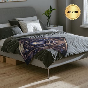 Pode incluir: Uma manta de arremesso cinza com um design de tape&ccedil;aria colorido que apresenta uma cena celestial com estrelas, luas e planetas. A tape&ccedil;aria est&aacute; dobrada em uma cama cinza com travesseiros brancos. O texto "The Magician" &eacute; vis&iacute;vel na tape&ccedil;aria.