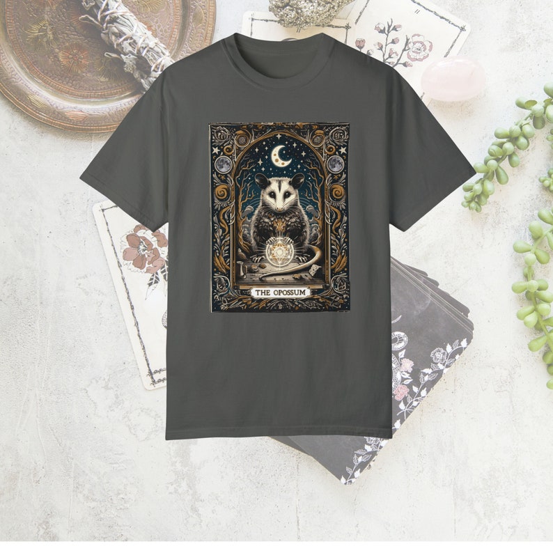 May include: A dark gray t-shirt with a graphic print of a possum holding a crystal ball. The graphic is surrounded by a gold frame and features a crescent moon and stars. The text "THE OPOSSUM" is printed below the graphic.