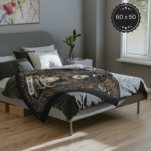 May include: A black throw blanket with a gold and white design featuring a tarot card with the text "The Possum". The blanket is on a grey bed with white pillows.