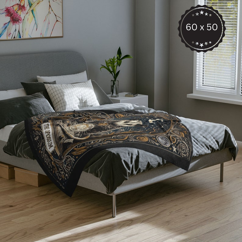 May include: A black throw blanket with a gold and white design featuring a tarot card with the text "The Possum". The blanket is on a grey bed with white pillows.