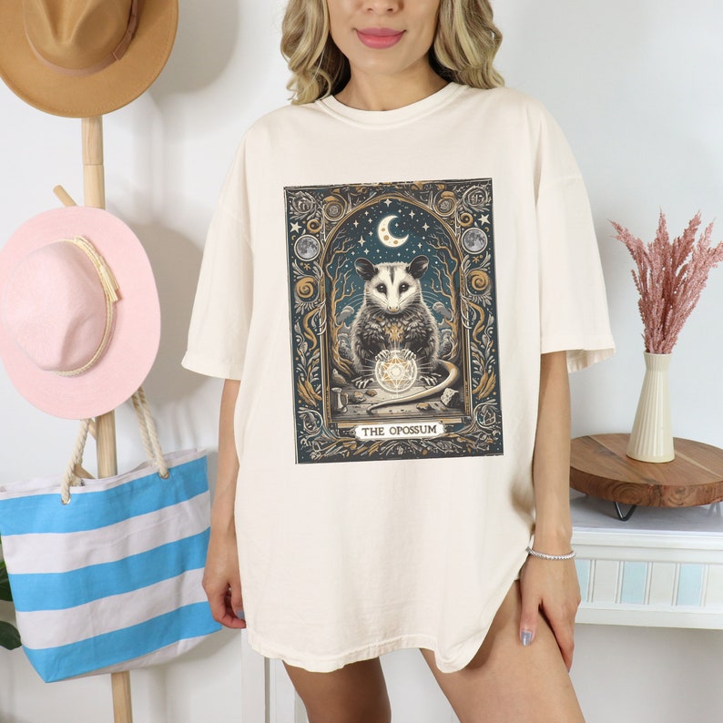 May include: A cream-colored oversized t-shirt with a graphic print of an opossum holding a crystal ball. The graphic is surrounded by a gold frame and features a crescent moon and stars. The text "THE OPOSSUM" is printed below the graphic.