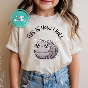 May include: Beige t-shirt with the text "THIS IS HOW I ROLL" above a cartoon pill bug illustration. The pill bug is gray with a cute face and sparkles. The shirt is worn by a child.
