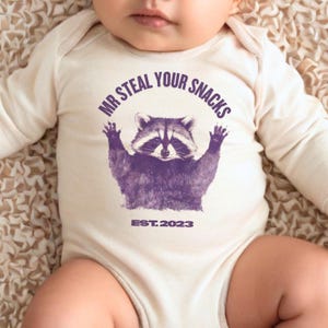 Pu&ograve; includere: Un body da bambino color crema a maniche lunghe. Il body presenta una grafica viola di un procione con le braccia alzate e la scritta "MR STEAL YOUR SNACKS" sopra il procione. Sotto il procione c'&egrave; la scritta "EST. 2023".