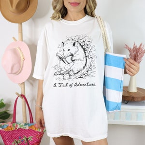Puede incluir: Camiseta blanca de gran tama&ntilde;o con una ilustraci&oacute;n en blanco y negro de un opossum leyendo un libro. El texto "A Tail of Adventure" est&aacute; impreso debajo de la ilustraci&oacute;n.