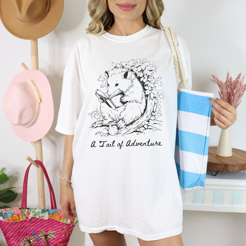Puede incluir: Camiseta blanca de gran tama&ntilde;o con una ilustraci&oacute;n en blanco y negro de un opossum leyendo un libro. El texto "A Tail of Adventure" est&aacute; impreso debajo de la ilustraci&oacute;n.