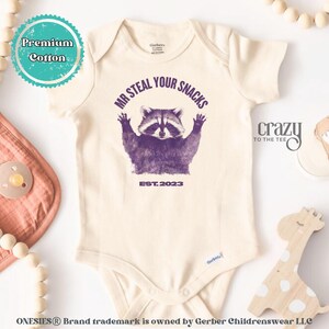 Pu&ograve; includere: Body da bambino color crema con grafica di procione viola e la scritta "MR STEAL YOUR SNACKS EST. 2023". Il body ha maniche corte e chiusura con bottoni automatici. Le parole "Premium Cotton" sono in un cerchio turchese.