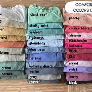 May include: A collection of 20 different colored t-shirts stacked in three columns. The t-shirts are labeled with names like "white", "ivory", "butter", "island reef", "chalky mint", "seafoam", "peachy", "crunch berry", "blossom", "mustard", "yam", "hydrange", "Ghambray", "watermelon", "heliconia", "blue jean", "sandstone", "Brick", "bay", "flo blue", "Chili", "orchid", "light green", "washed denim", "violet", "denim", "blue spruce", "mess", "gray", "berry", "pepper", and "black".