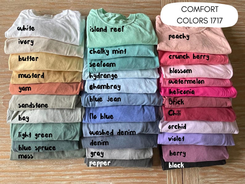 May include: A collection of 20 different colored t-shirts stacked in three columns. The t-shirts are labeled with names like "white", "ivory", "butter", "island reef", "chalky mint", "seafoam", "peachy", "crunch berry", "blossom", "mustard", "yam", "hydrange", "Ghambray", "watermelon", "heliconia", "blue jean", "sandstone", "Brick", "bay", "flo blue", "Chili", "orchid", "light green", "washed denim", "violet", "denim", "blue spruce", "mess", "gray", "berry", "pepper", and "black".