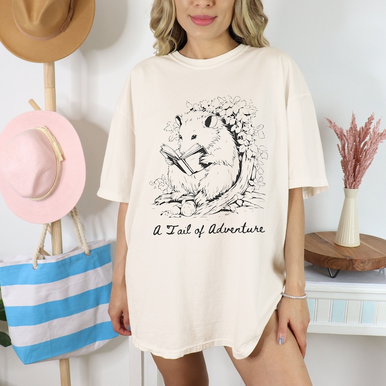 Puede incluir: Una camiseta de color crema con una ilustraci&oacute;n en blanco y negro de un opossum leyendo un libro. El texto "A Tail of Adventure" est&aacute; impreso debajo de la ilustraci&oacute;n.