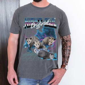 May include: A dark gray t-shirt with a retro-style graphic of opossums in a teacup. The graphic includes the text "Night Owl Original" in a colorful, stylized font.