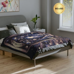 Pode incluir: Uma manta de arremesso azul escuro e dourada com um design celestial que apresenta estrelas, luas e o texto "The Raccoon".
