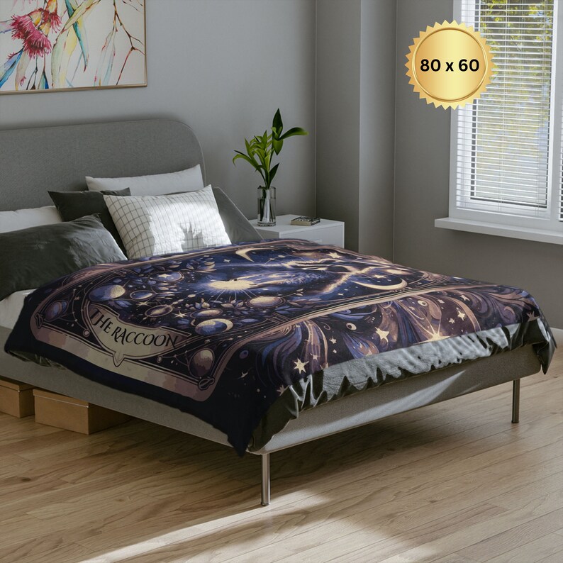 Pode incluir: Uma manta de arremesso azul escuro e dourada com um design celestial que apresenta estrelas, luas e o texto "The Raccoon".