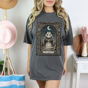 May include: A dark gray t-shirt with a graphic print of an opossum holding a crystal ball. The opossum is surrounded by a moon, stars, and other celestial imagery. The text "THE OPOSSUM" is printed below the image.