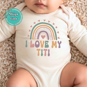 May include: A cream-colored baby onesie with long sleeves. The onesie features a rainbow design in pastel colors with the text "I LOVE MY TITI". Small hearts are above the rainbow. A turquoise circle with the words "Ships Fast" is in the upper left corner.