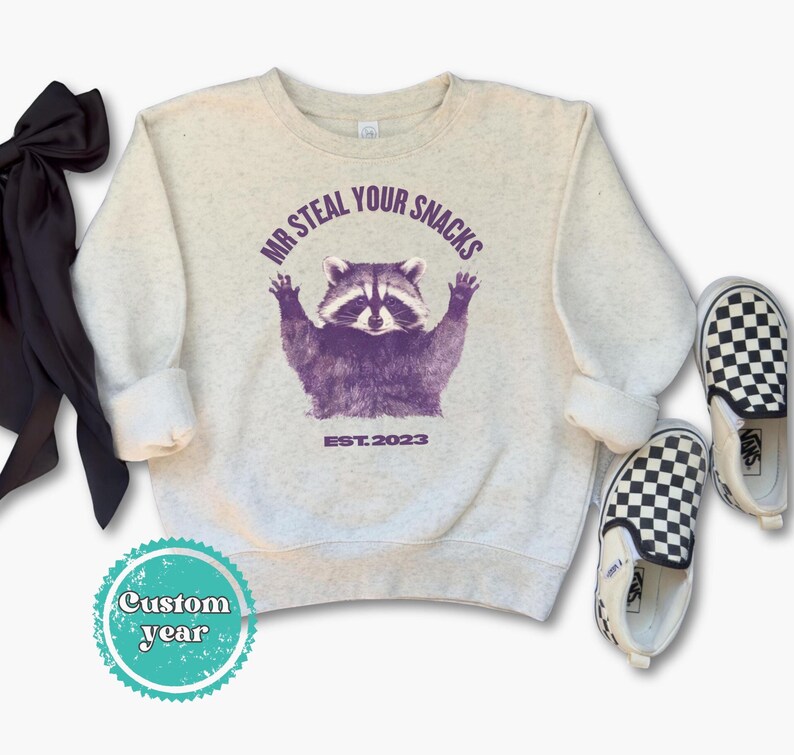 Pu&ograve; includere: Una felpa grigio chiaro con una grafica di procione viola e la scritta "MR STEAL YOUR SNACKS EST.2023". L'immagine include anche scarpe slip-on a scacchi bianchi e neri e un fiocco nero.